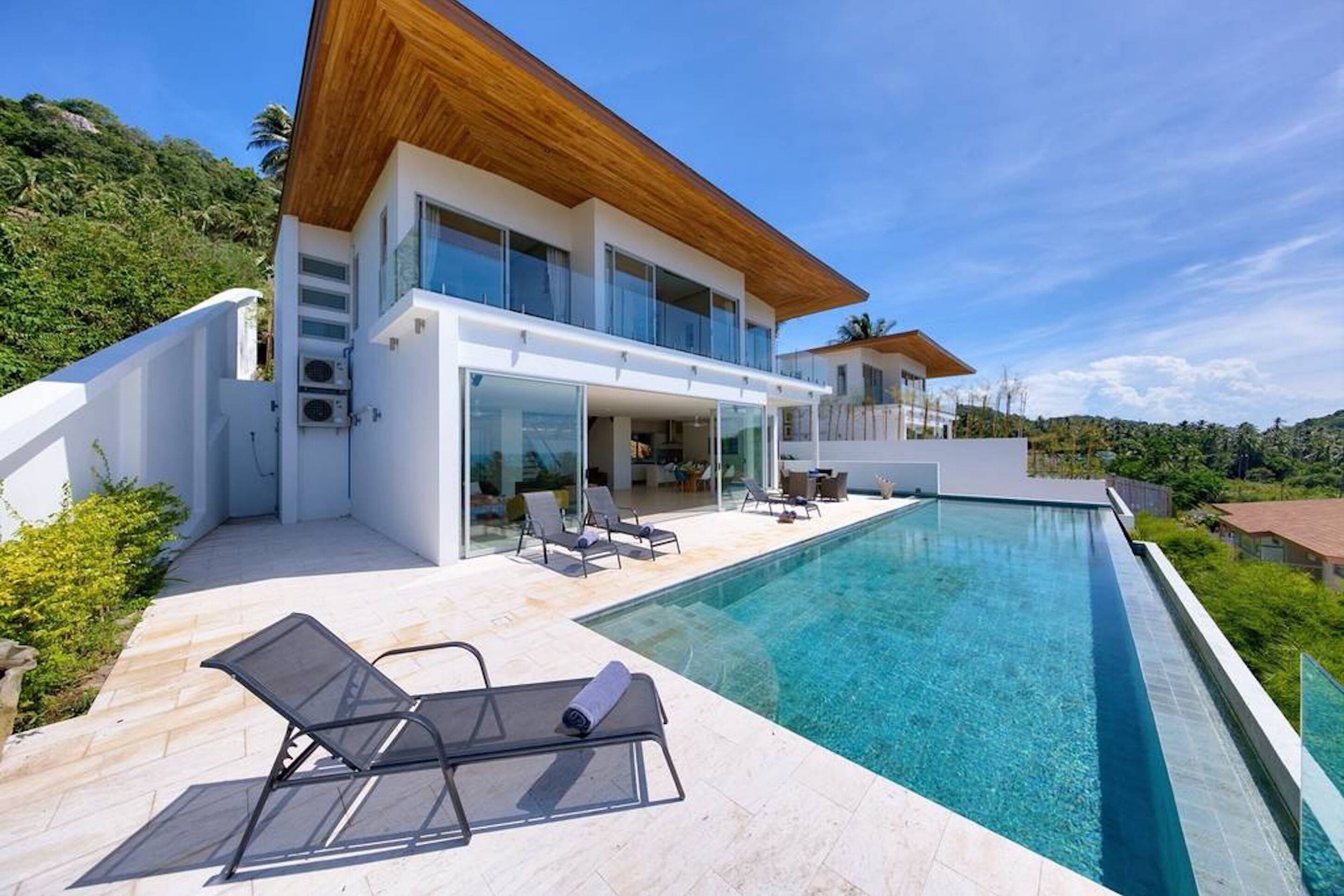 Une maison moderne dotée d’une grande piscine, d’un espace salon extérieur et offrant une vue sur la verdure environnante.