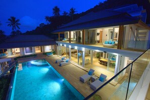 Una casa moderna con una gran piscina, un balcón en el segundo piso con asientos y un interior bien iluminado.