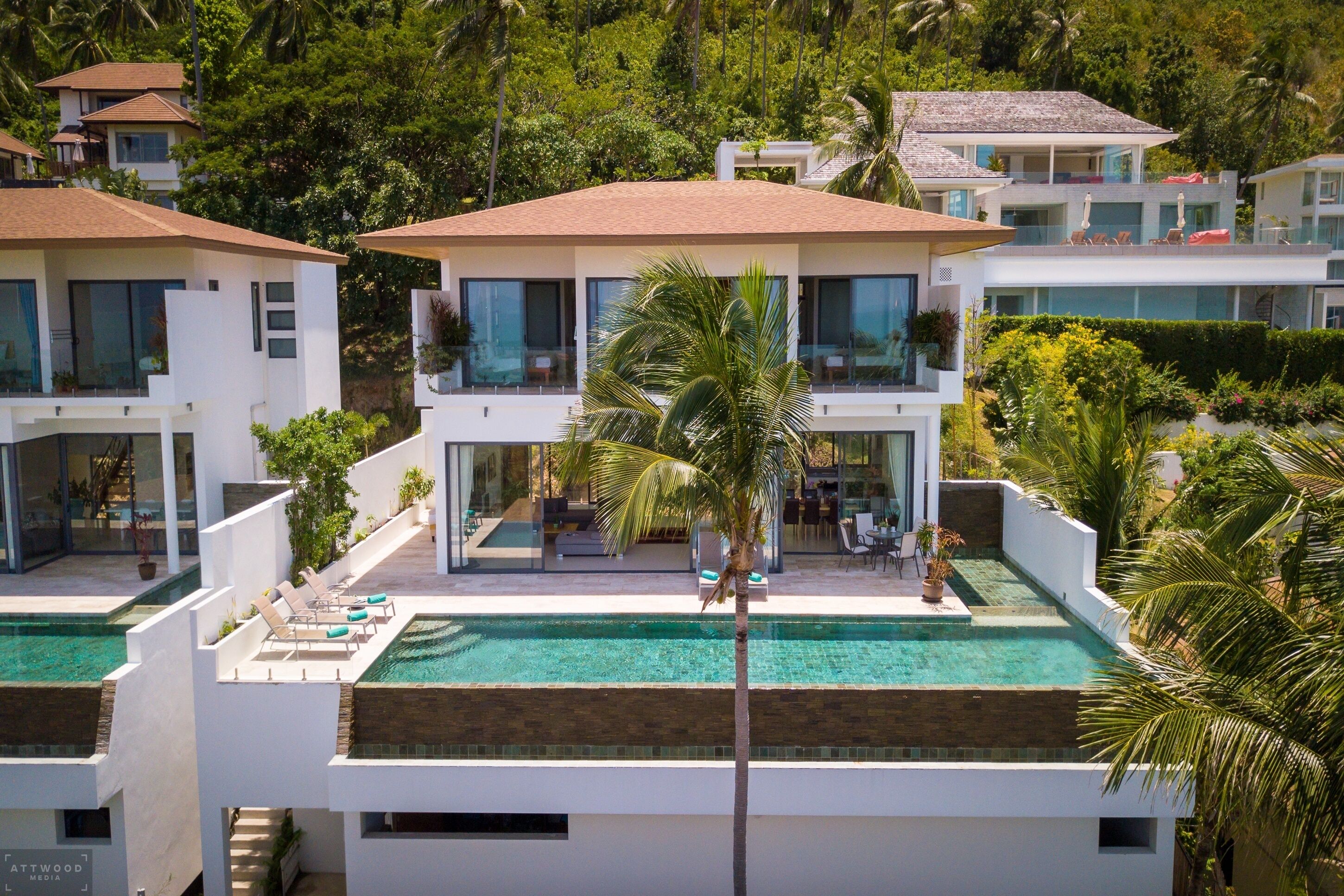 Une maison moderne à plusieurs étages, dotée d’une piscine sur le toit, entourée d’une végétation luxuriante et de palmiers.