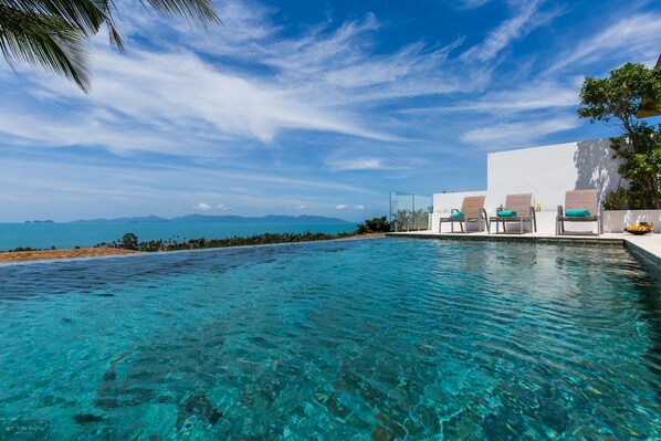A clear blue pool with lounge chairs and a white building with a view of the ocean and mountains.