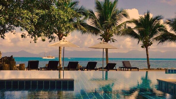 Una piscina infinita con sillones de descanso y sombrillas, con vista a una playa con palmeras y un barco a lo lejos.