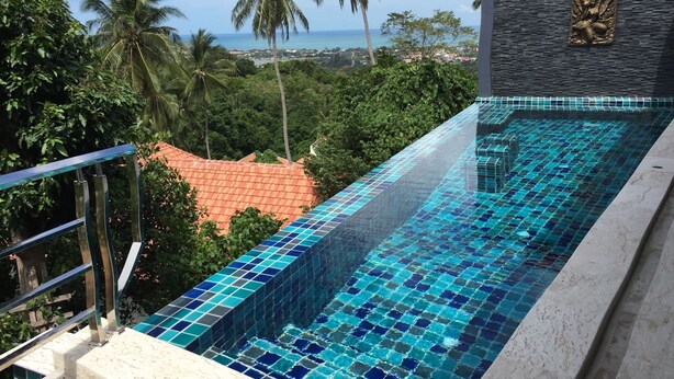 Una piscina con piastrelle di mosaico blu, immersa nel verde e con vista sull'oceano.