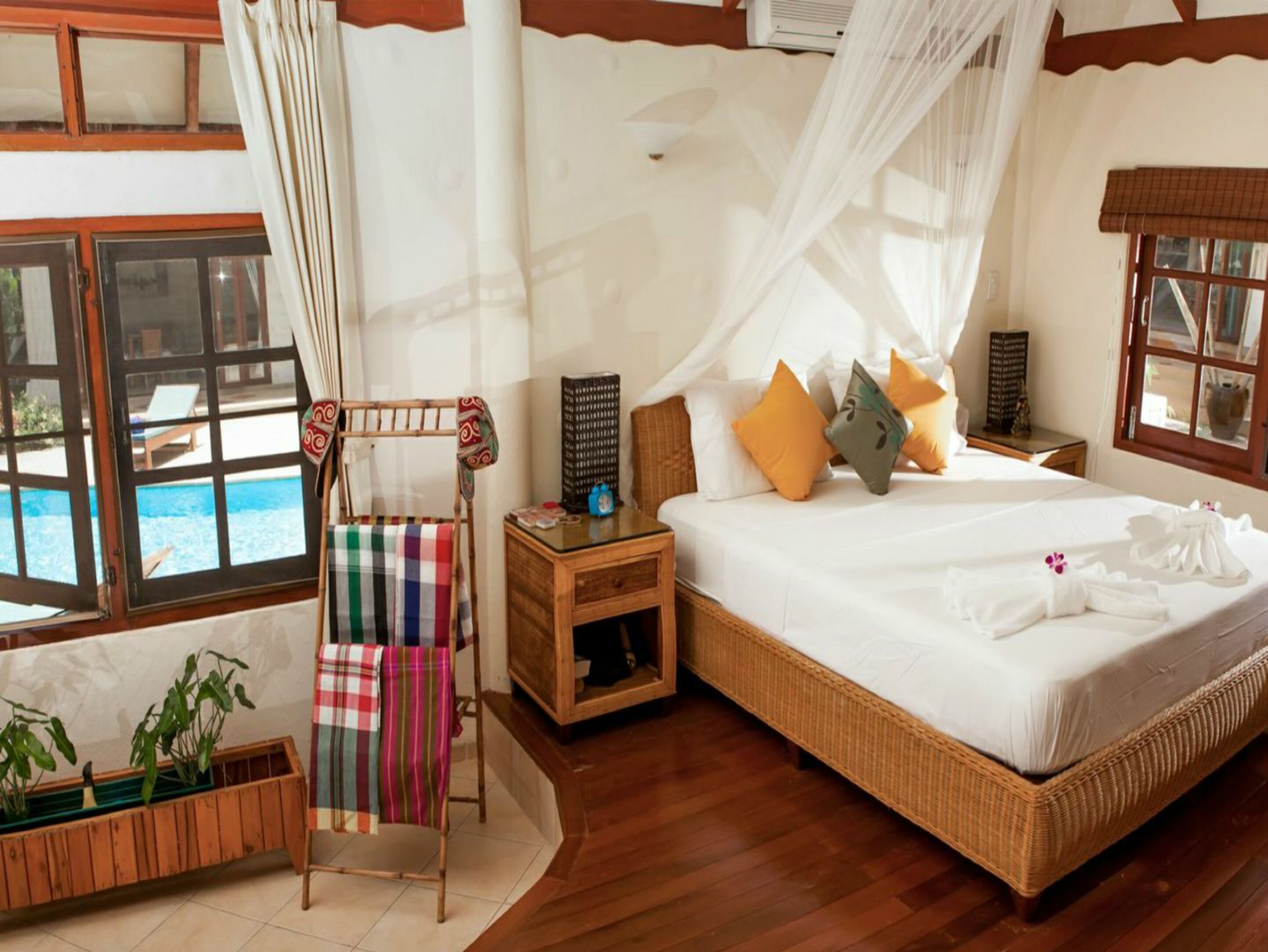 A bedroom with a large bed, a bedside table, a nightstand, a wooden chair with a colorful blanket, a small table with a decorative item, and a window with a view of a pool.