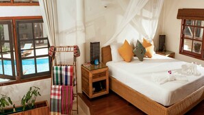 Een slaapkamer met een groot bed, een nachtkastje, een houten stoel met een kleurrijke deken, een klein tafeltje met een decoratief object, en een raam met uitzicht op een zwembad.