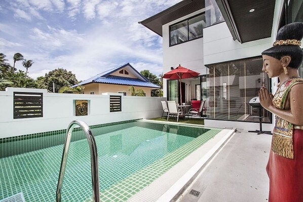 Una piscina con escalera metálica, una casa moderna con tejado azul y una estatua de una mujer con traje tradicional.
