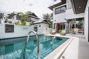 Una piscina con escalera metálica, rodeada por un muro blanco con diseño de azulejos. Hay dos sillas de descanso y una mesa con sillas fuera de la zona de la piscina.