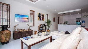 A living room with a sofa, coffee table, and decorative items. There is a TV, artwork, and a kitchen in the background. - 6 Bedroom Twin Villas Ban Tai near Beach SDV260/261-By Samui Dream Villas (Koh Samui)