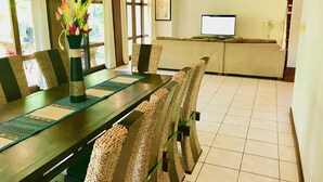 Un comedor con una mesa larga, sillas, un florero con flores y un televisor al fondo.