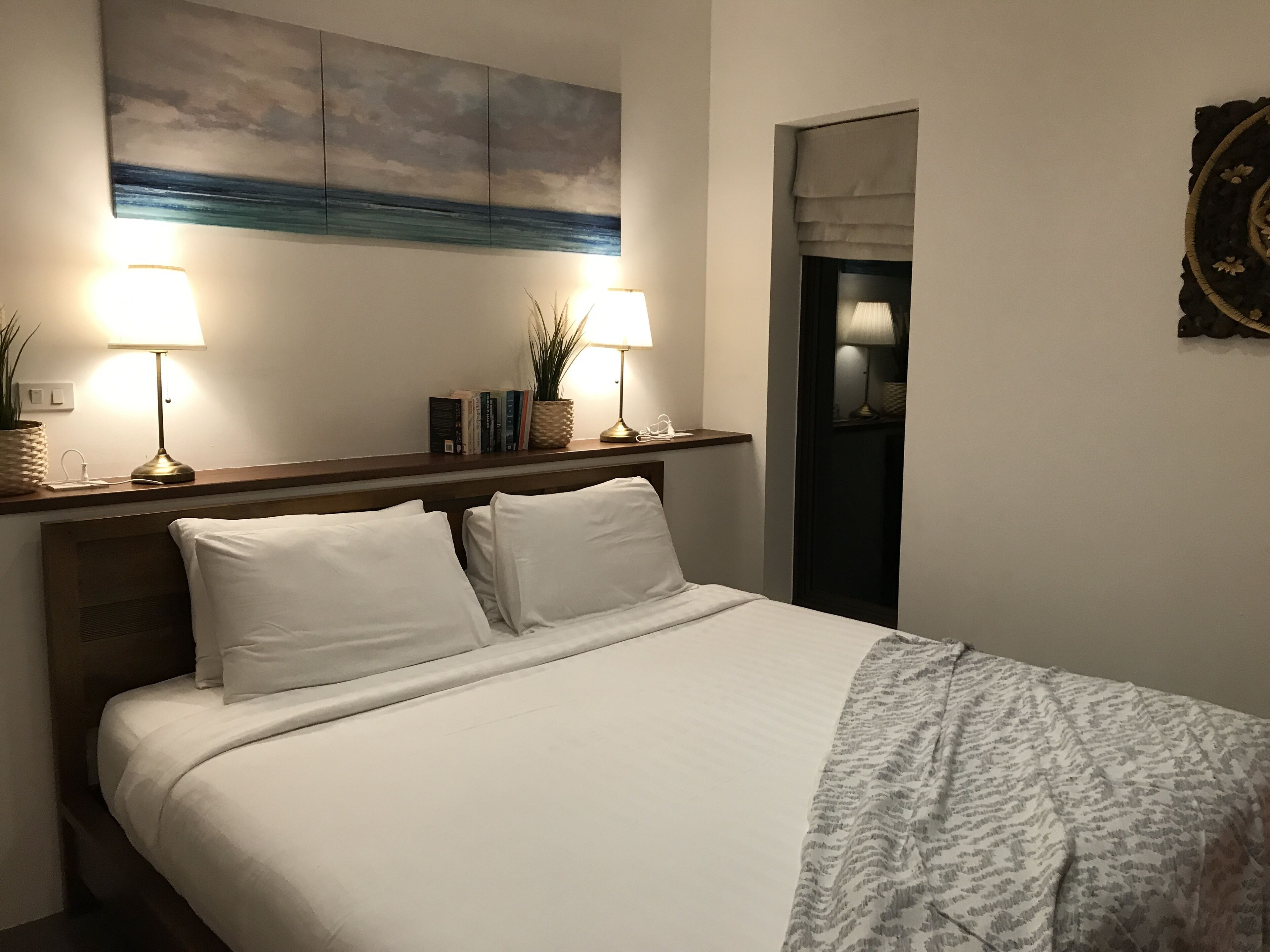 A neatly made bed with white linens, two bedside lamps, a shelf with books and a plant, and a painting above the bed.