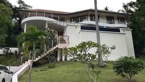 A modern two-story house with a curved balcony, a stone retaining wall, and a small garden with palm trees. - 3 Bedroom Sea View Villa Santi SDV228-By Samui Dream Villas (Koh Samui)