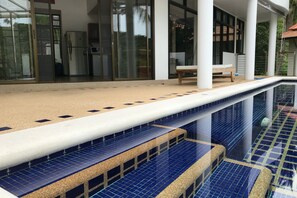 Zwembad met blauwe tegels en een witte rand, omgeven door een overdekt terras met een bank en een koelkast.