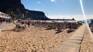 Uma praia com espreguiçadeiras e guarda-sóis, um calçadão de madeira e um penhasco ao fundo.