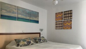 Cama bem arrumada com edredom branco e duas almofadas com padrão de folhas, cabeceira de madeira e parede com três quadros e quatro painéis decorativos estampados.