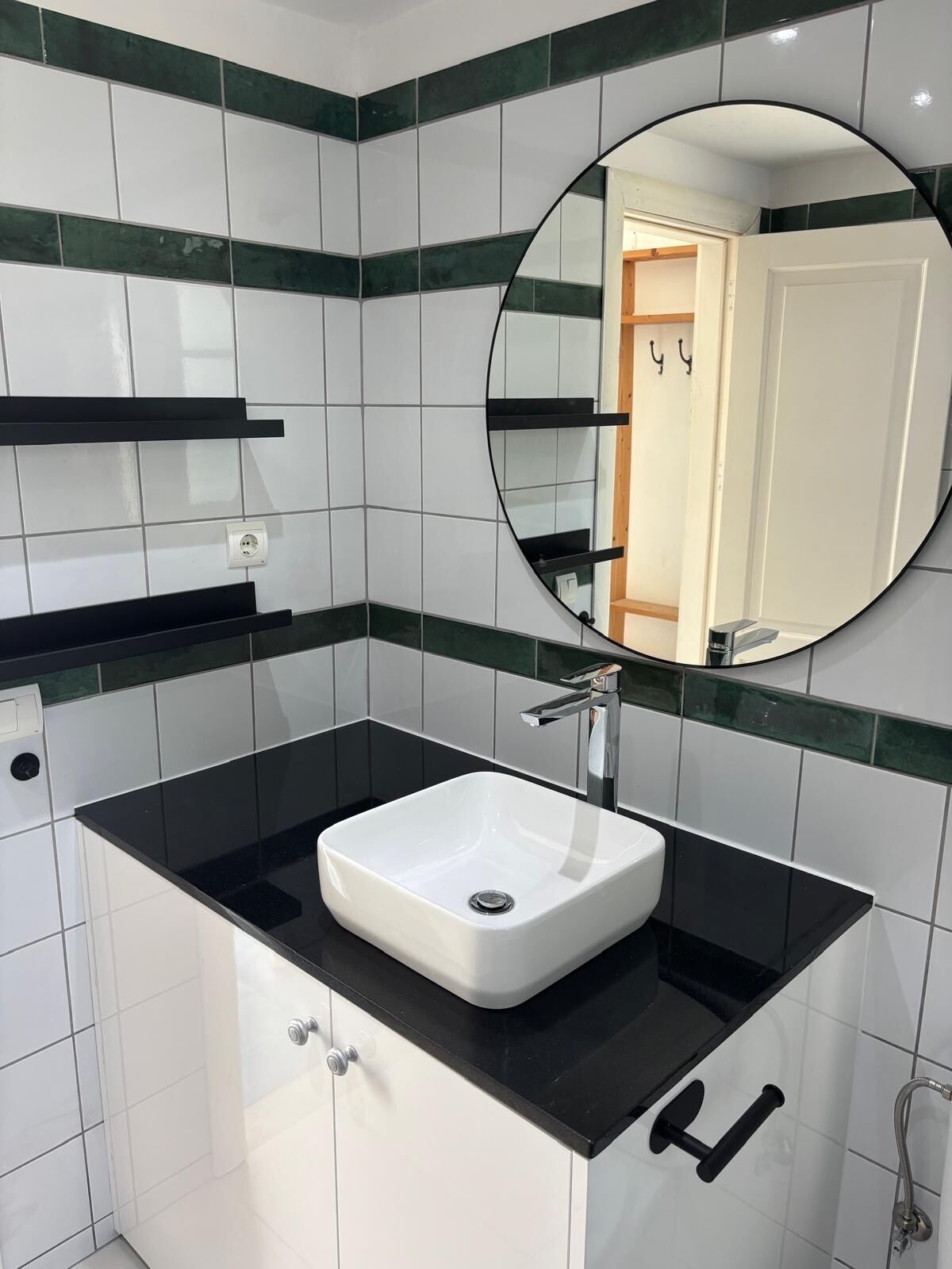A modern bathroom with a white sink, black countertop, and green-tiled walls. There is a mirror above the sink and a shower area to the right.
