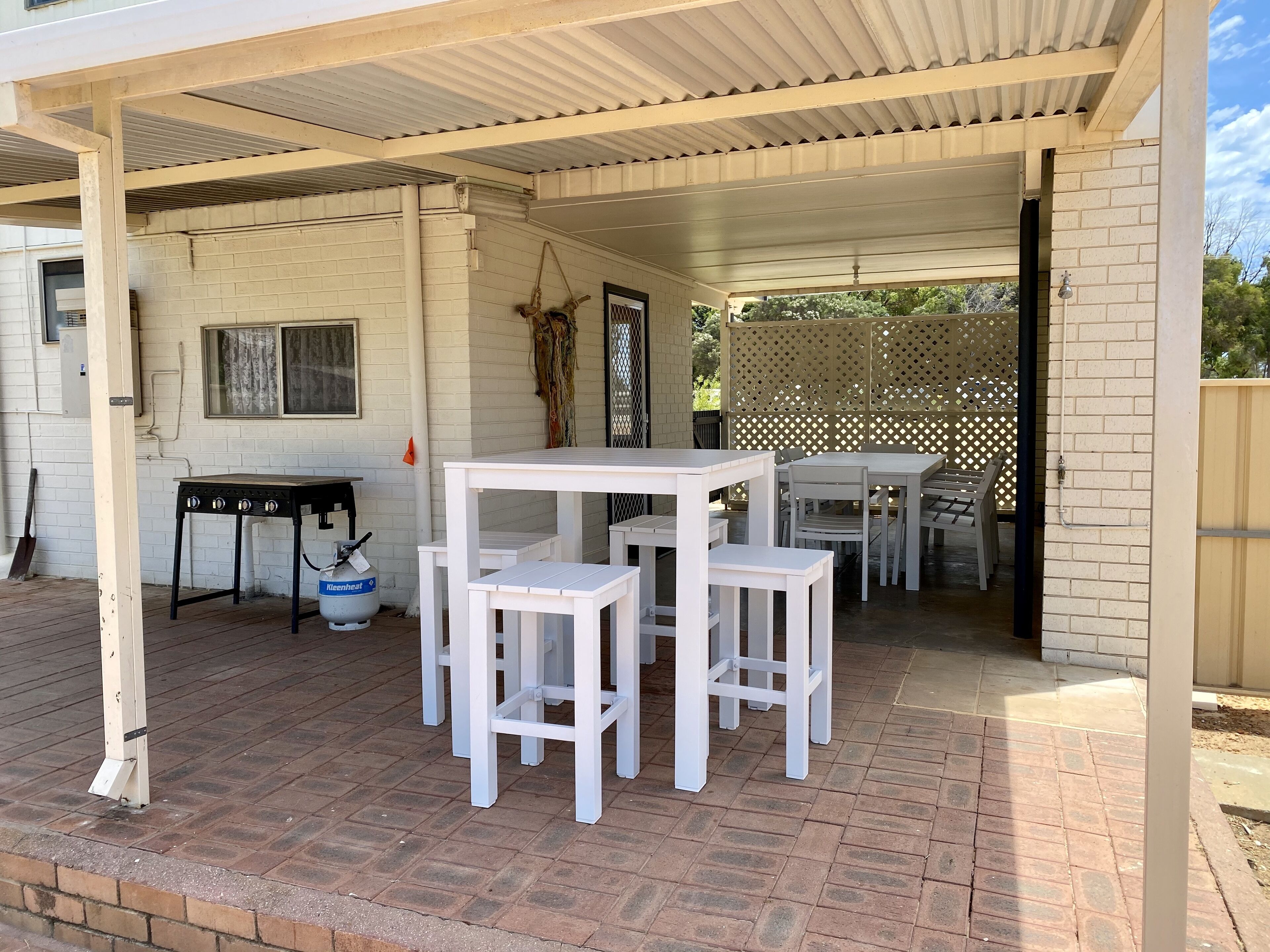 A covered patio area with white tables and chairs, a gas cylinder, and a brick wall.