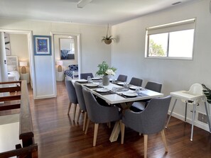 A dining area with a long table set for six, surrounded by grey chairs. There is a white chair to the right, and a small white table with a plant. The room has wooden flooring and white walls. - Location Is Everything plus WiFi (Jurien Bay)