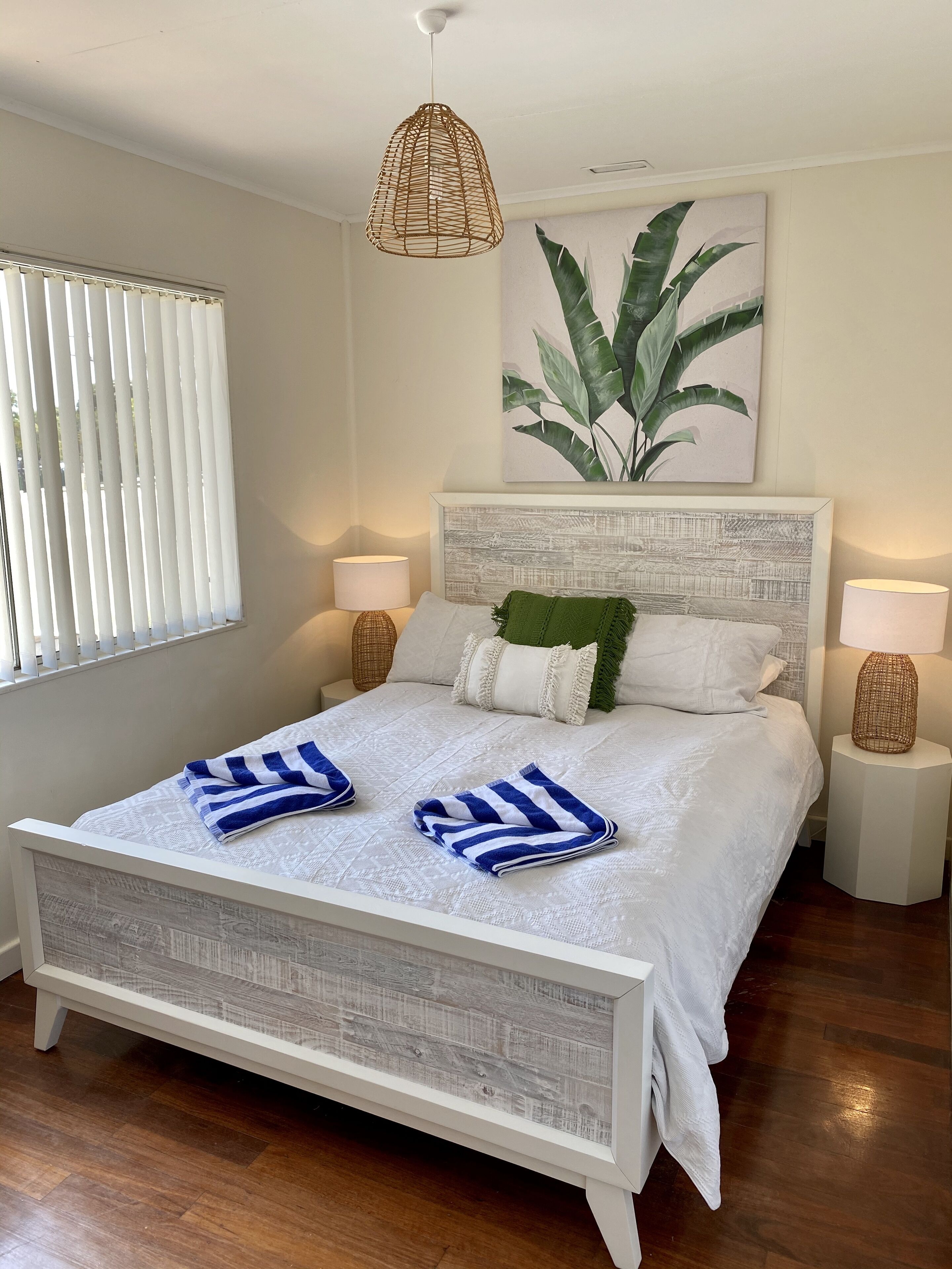 A neatly made bed with white bedding and blue striped towels, a woven pendant light, a framed botanical print, and two bedside lamps.