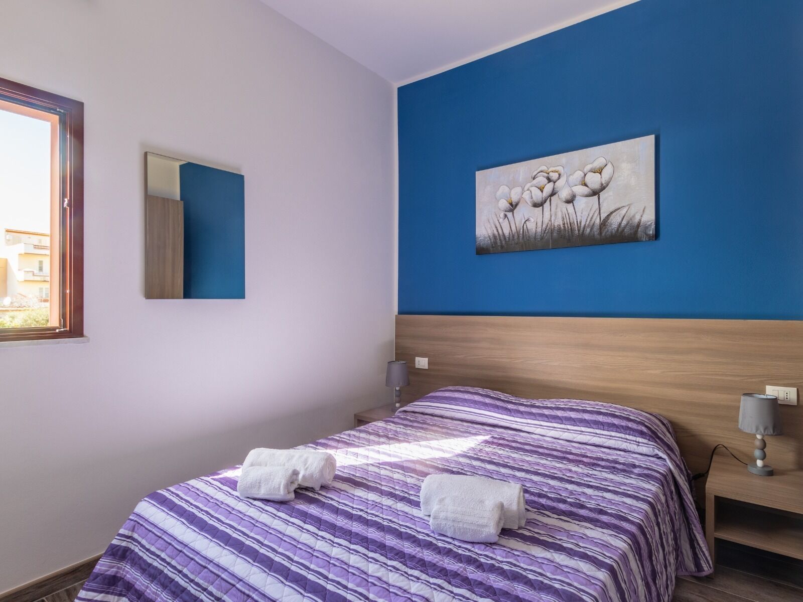 A bedroom with a bed, a painting, a window, and a bedside table.