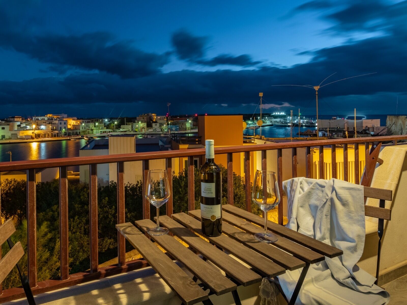 Una mesa de madera con una botella de vino y copas en un balcón con vistas a un puerto deportivo con barcos y edificios.