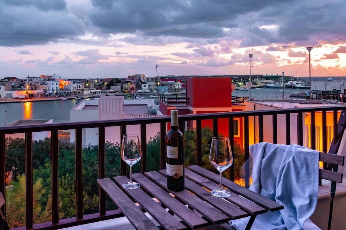 A balcony with a wooden table, two wine glasses, and a bottle of wine.
