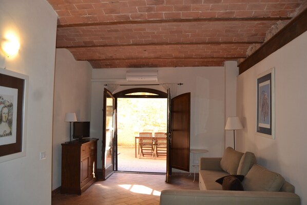 A living room with a sofa, a TV on a wooden cabinet, and an open door leading to an outdoor area with a table and chairs.