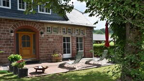 A brick house with a wooden front door, a patio with two lounge chairs, and a red umbrella. - 90m² holiday house in Dhronecken (Dhronecken)