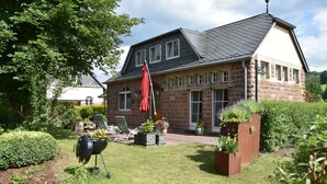 A two-story stone house with a sloped roof, a patio with outdoor furniture, and a well-maintained garden. - 90m² holiday house in Dhronecken (Dhronecken)