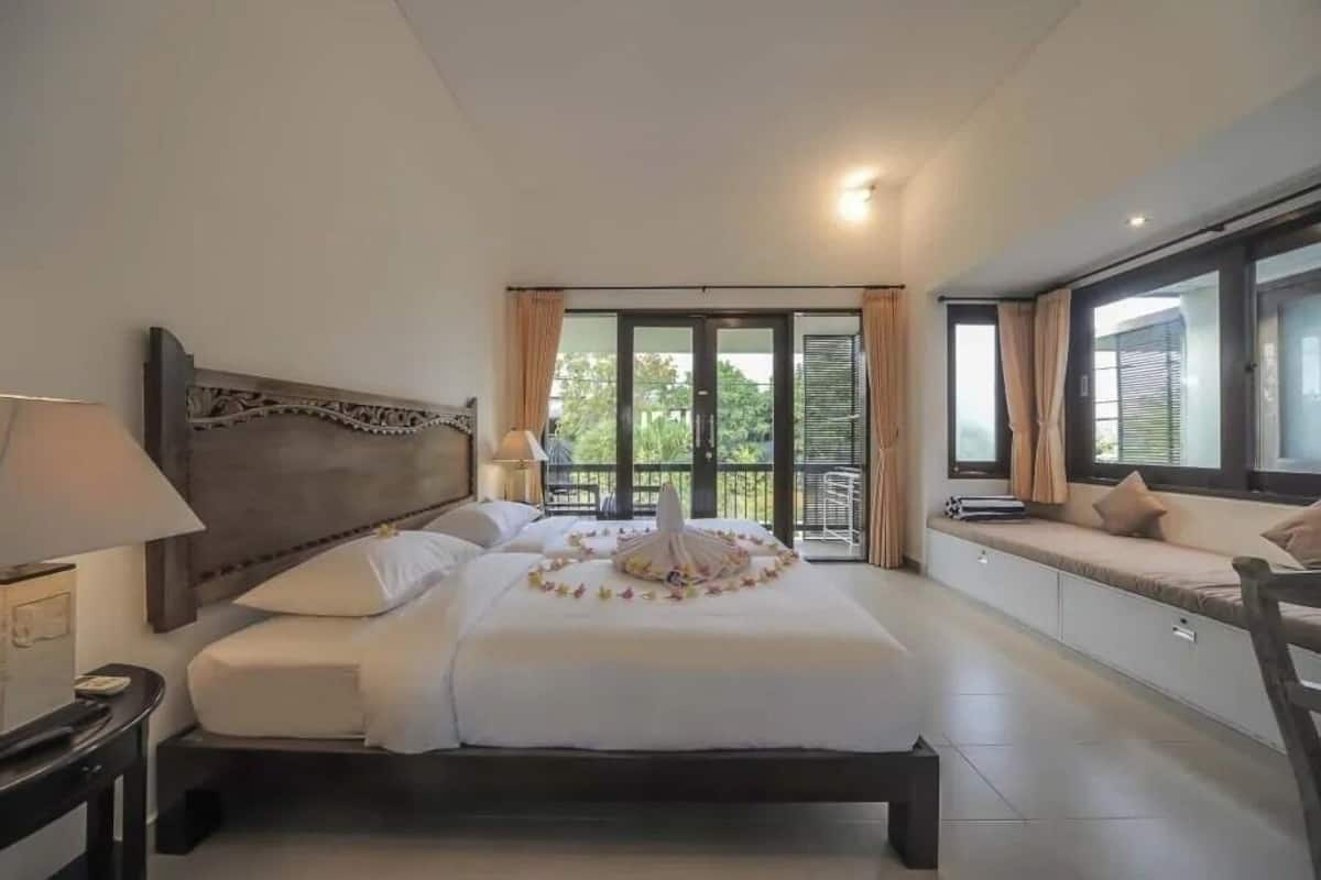 A hotel room with a large bed, a bedside table, a lamp, a window with curtains, and a balcony with a seating area.