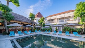 A swimming pool with lounge chairs and umbrellas, surrounded by buildings with a thatched roof and a red-roofed structure. - Sayang Sanur Resort (Denpasar)