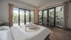 A hotel room with a large bed, a wooden bench, and a view of greenery through the sliding glass doors. - Sayang Sanur Resort (Denpasar)