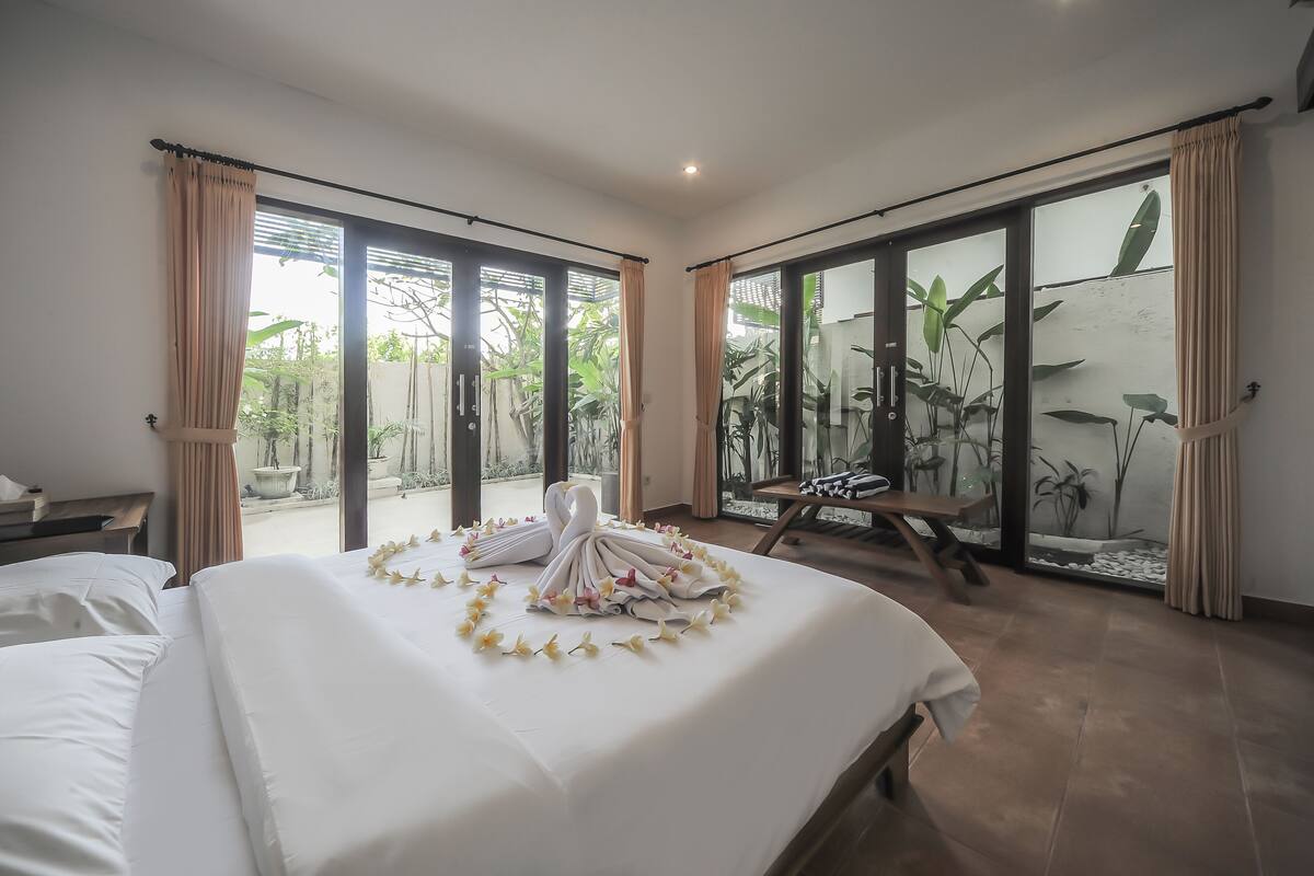 A hotel room with a large bed, a wooden bench, and a view of greenery through the sliding glass doors.