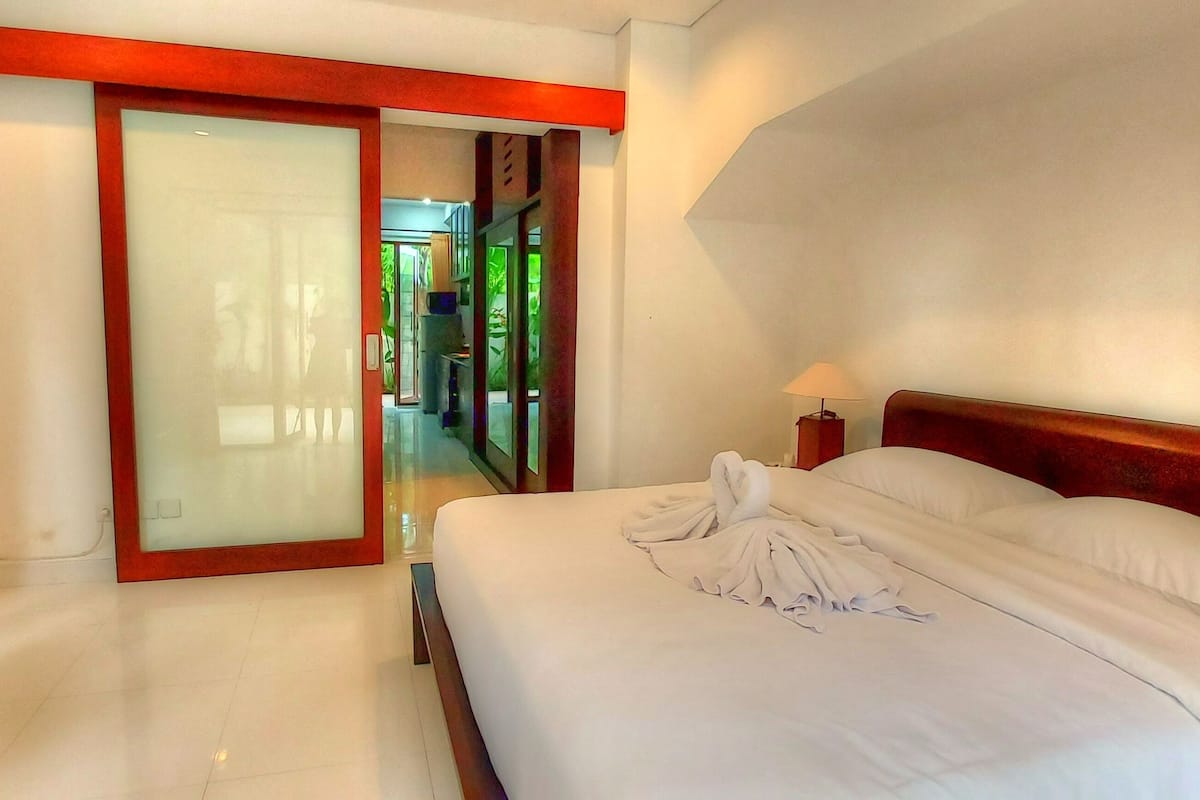 A hotel room with a bed, a nightstand, a door leading to a hallway, and a view of the hallway.