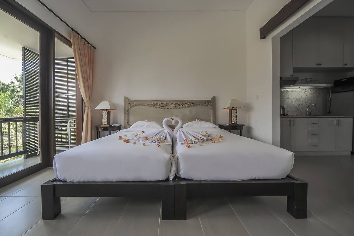 A double bed with a heart-shaped decoration on the pillows, a nightstand with a lamp, and a view of a balcony and kitchen area.