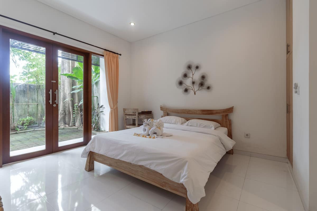 A bedroom with a wooden bed, white bedding, a decorative wall piece, and a sliding glass door leading to a garden.