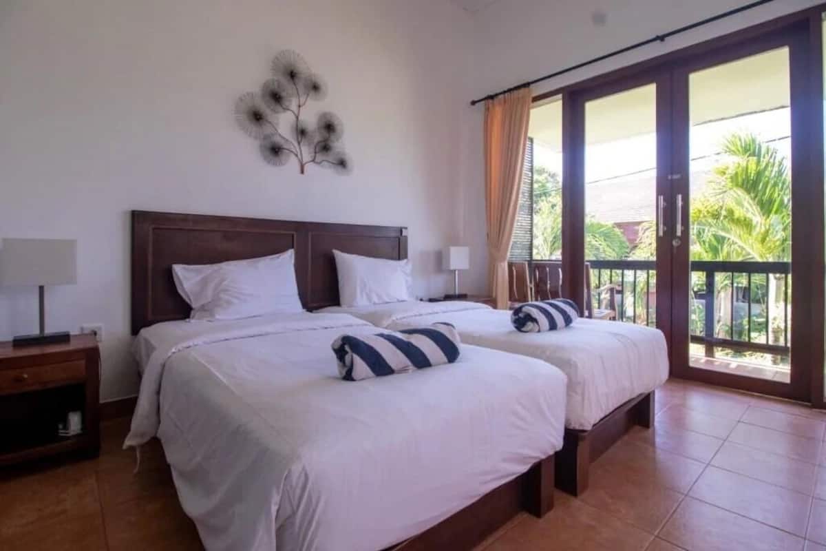 A hotel room with two beds, a wooden headboard, a nightstand with a lamp, and a balcony with curtains.