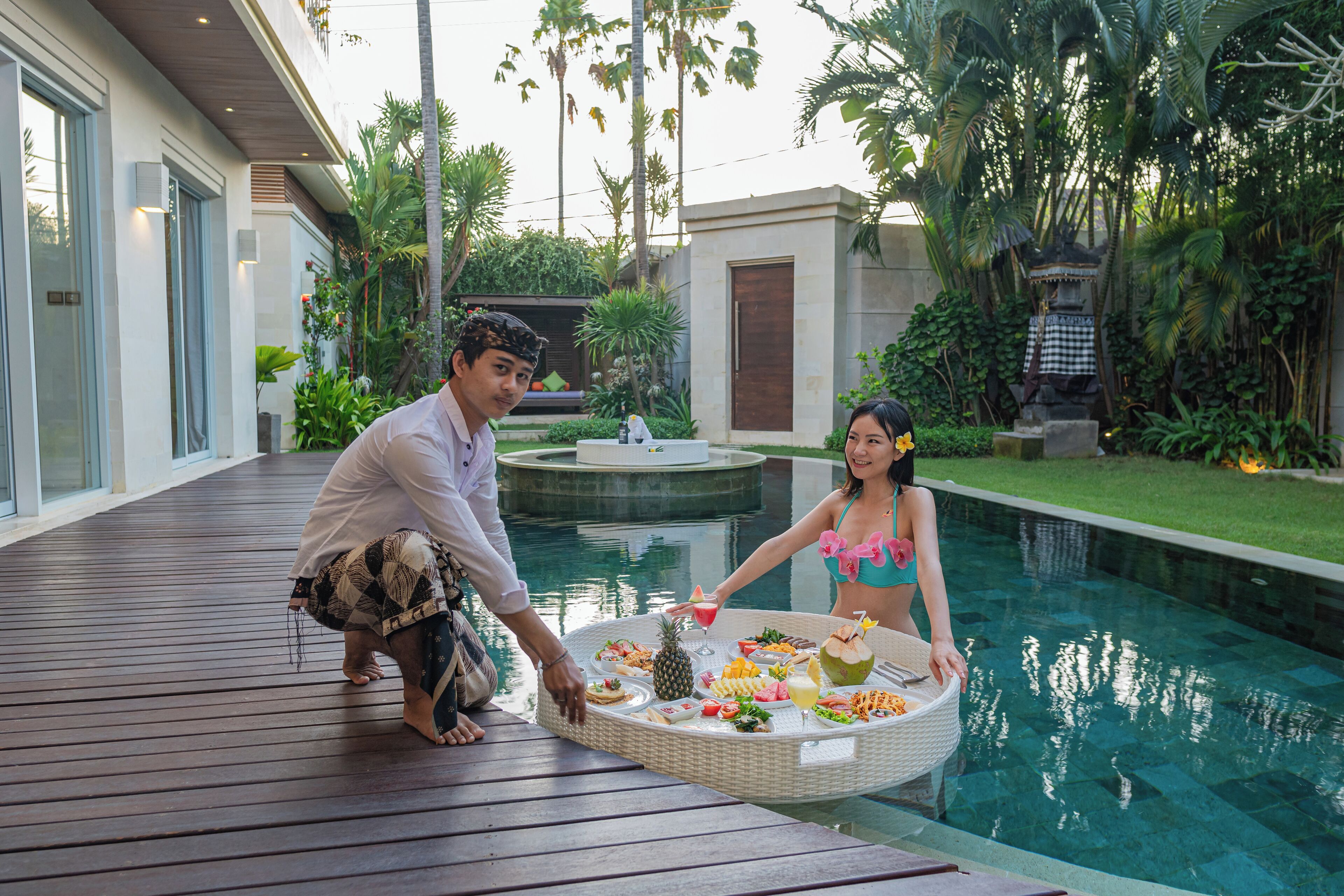 Um casal sentado à mesa junto à piscina, com comida e bebidas, em meio a vegetação exuberante e um edifício moderno.