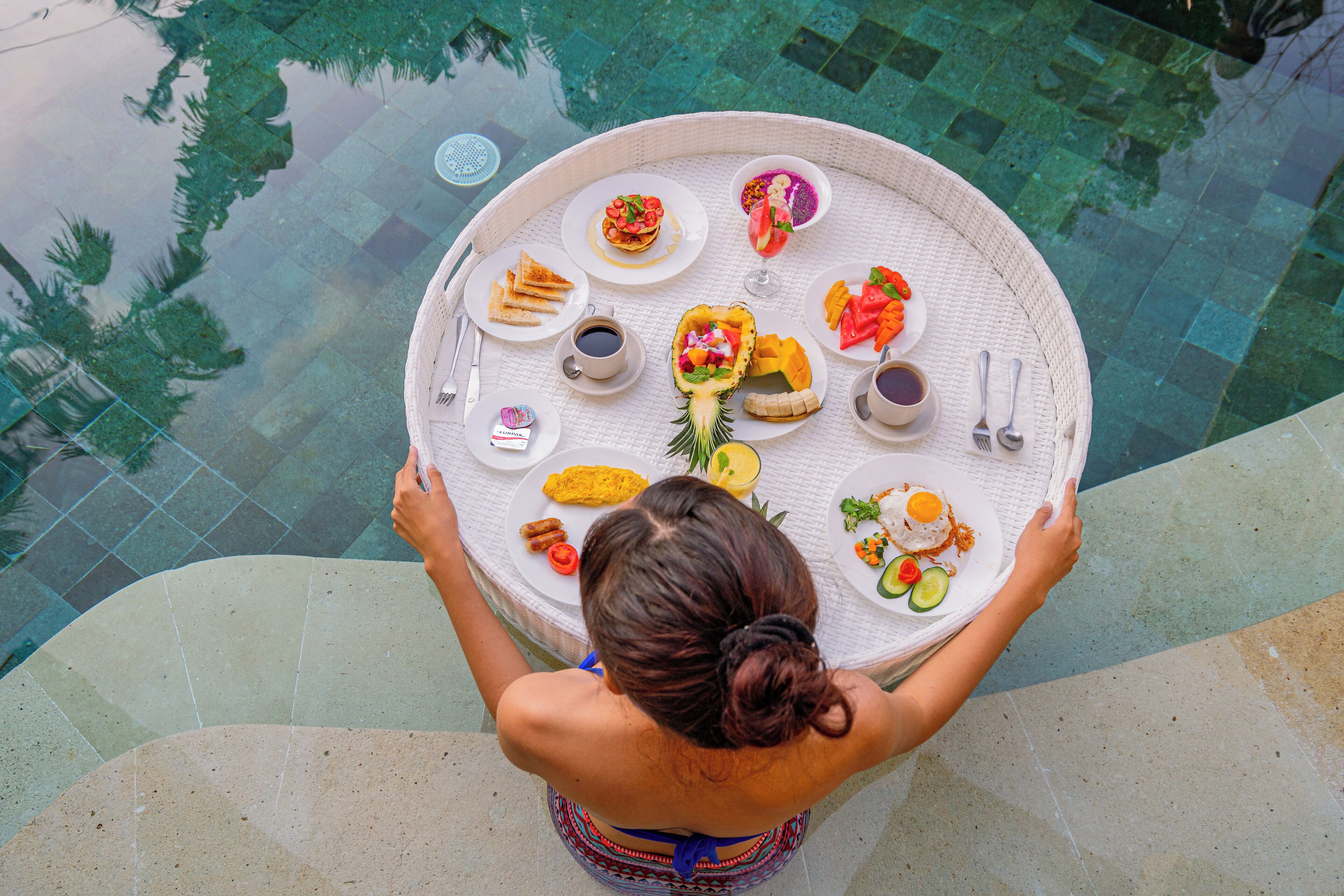 A round dining table with various dishes and drinks, held by a person standing by a pool.