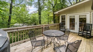 A wooden deck with a round table and chairs, a grill, and a view of a forested area. - Delightful Walk to Town Cottage in the Harris Lake Area | Cardinal Cottage (Highlands)