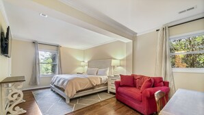 A bedroom with a bed, nightstands, a red sofa, a wooden table, and a TV. - Delightful Walk to Town Cottage in the Harris Lake Area | Cardinal Cottage (Highlands)