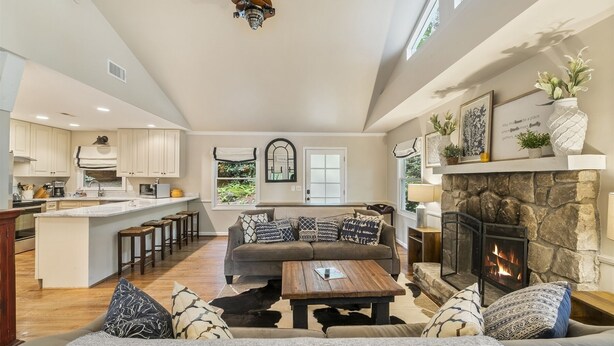 A spacious living area with a fireplace, a kitchen with a bar, and a large window overlooking greenery. - Delightful Walk to Town Cottage in the Harris Lake Area | Cardinal Cottage (Highlands)