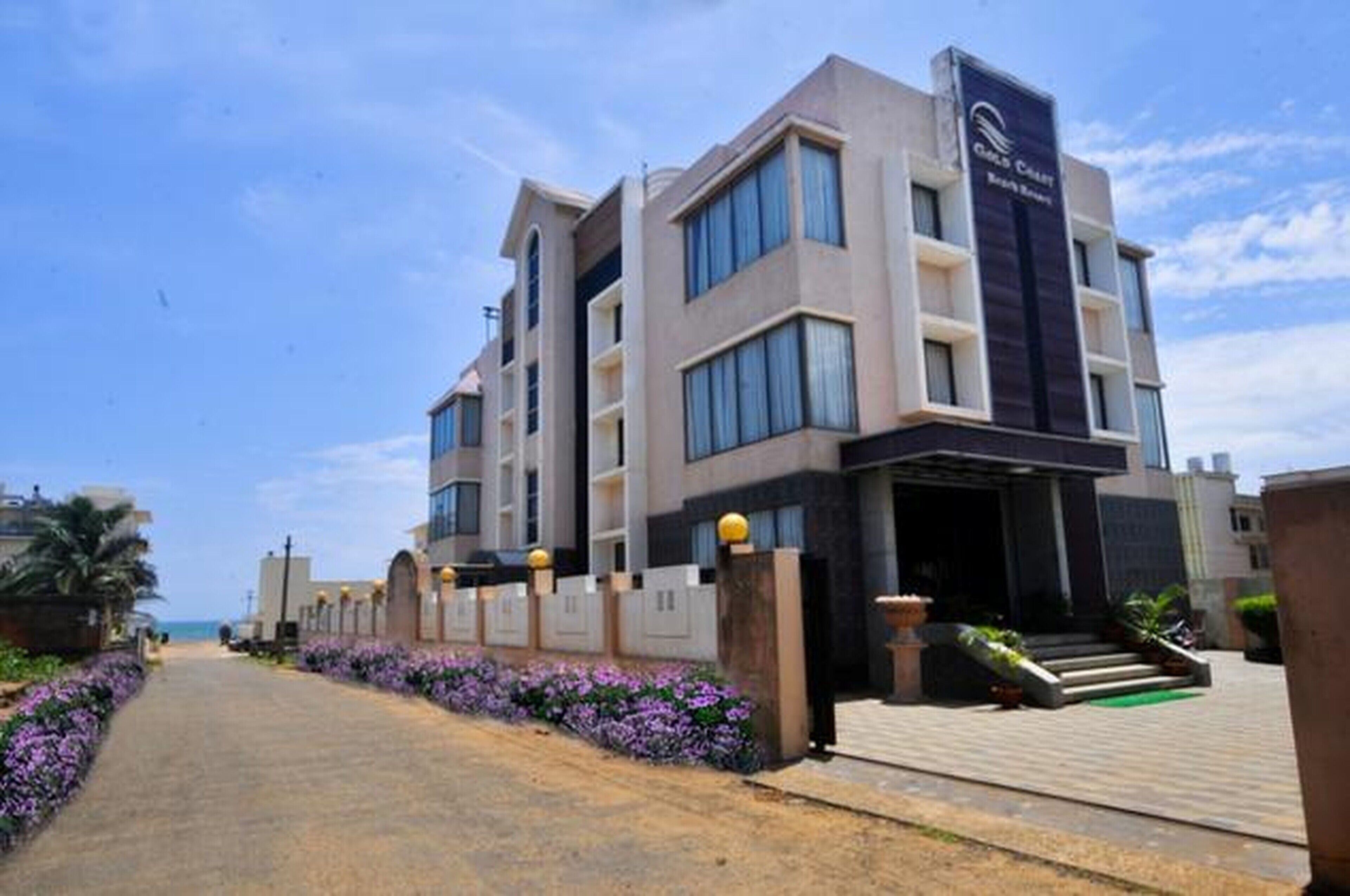 A modern hotel with a prominent entrance, a pathway lined with flowers, and a clear view of the sea in the background.