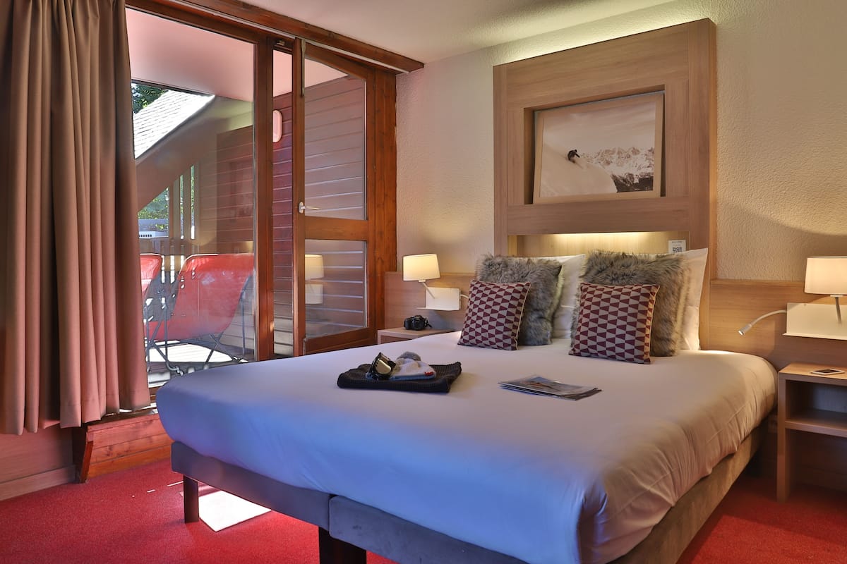 Ein Hotelzimmer mit einem großen Bett, einem Holzkopfteil, einem gerahmten Bild und einem Balkon mit roten Stühlen.