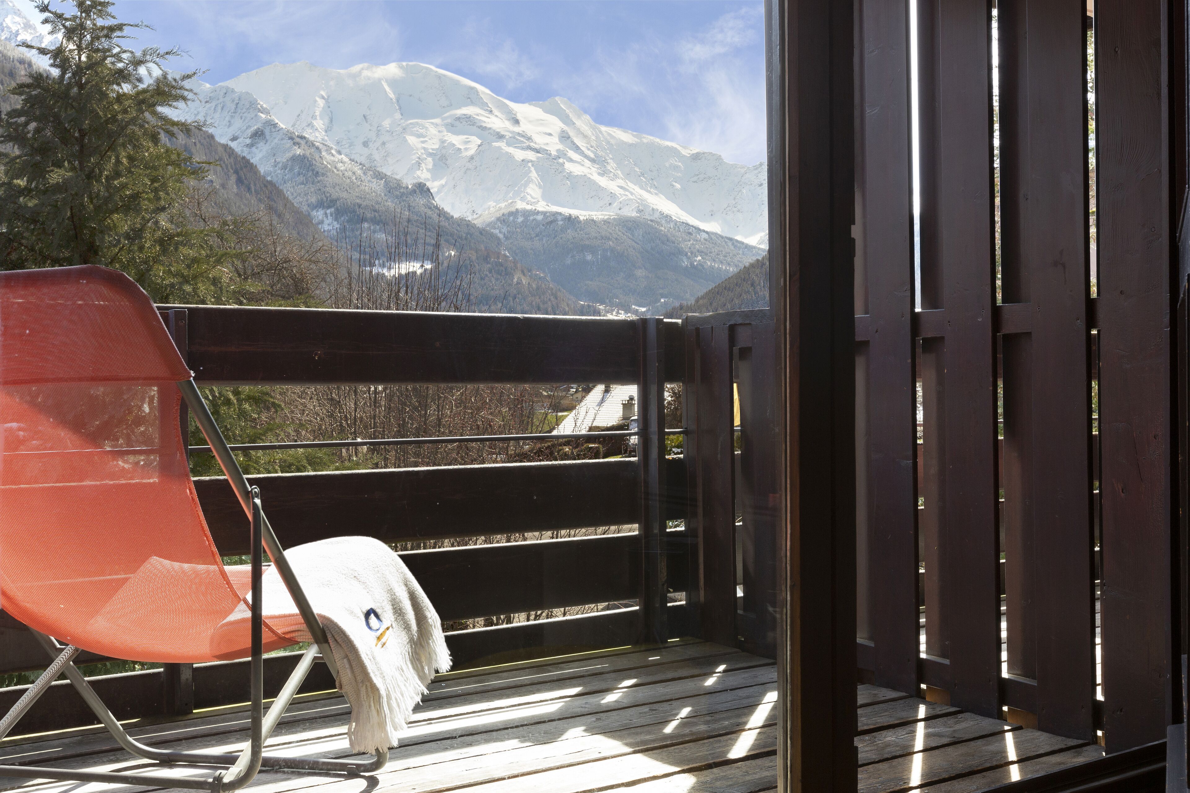 A wooden deck with a red chair and a white towel, overlooking a snow-capped mountain.