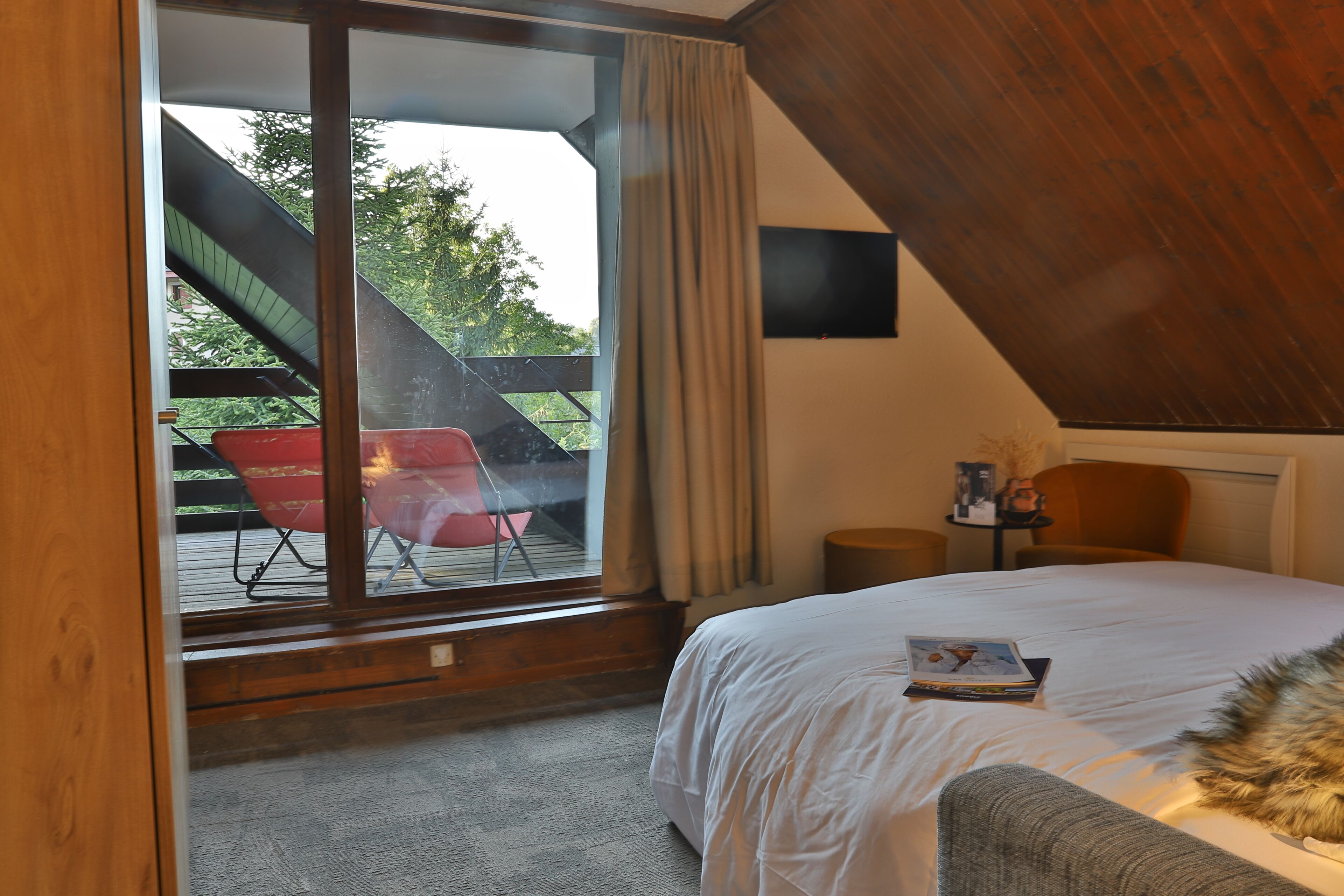 Une chambre avec un grand lit, une fenêtre avec des rideaux, un plafond en bois et une vue sur un espace extérieur où se trouve une chaise rouge.