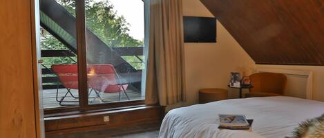 Une chambre avec un grand lit, une fenêtre avec des rideaux, un plafond en bois et une vue sur un espace extérieur où se trouve une chaise rouge.