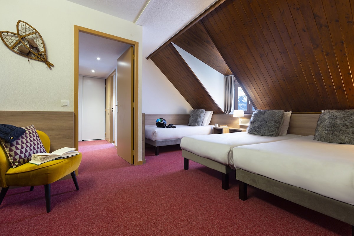 Ein Zimmer mit zwei Betten, einem gelben Sessel und einer Holzbalkendecke.