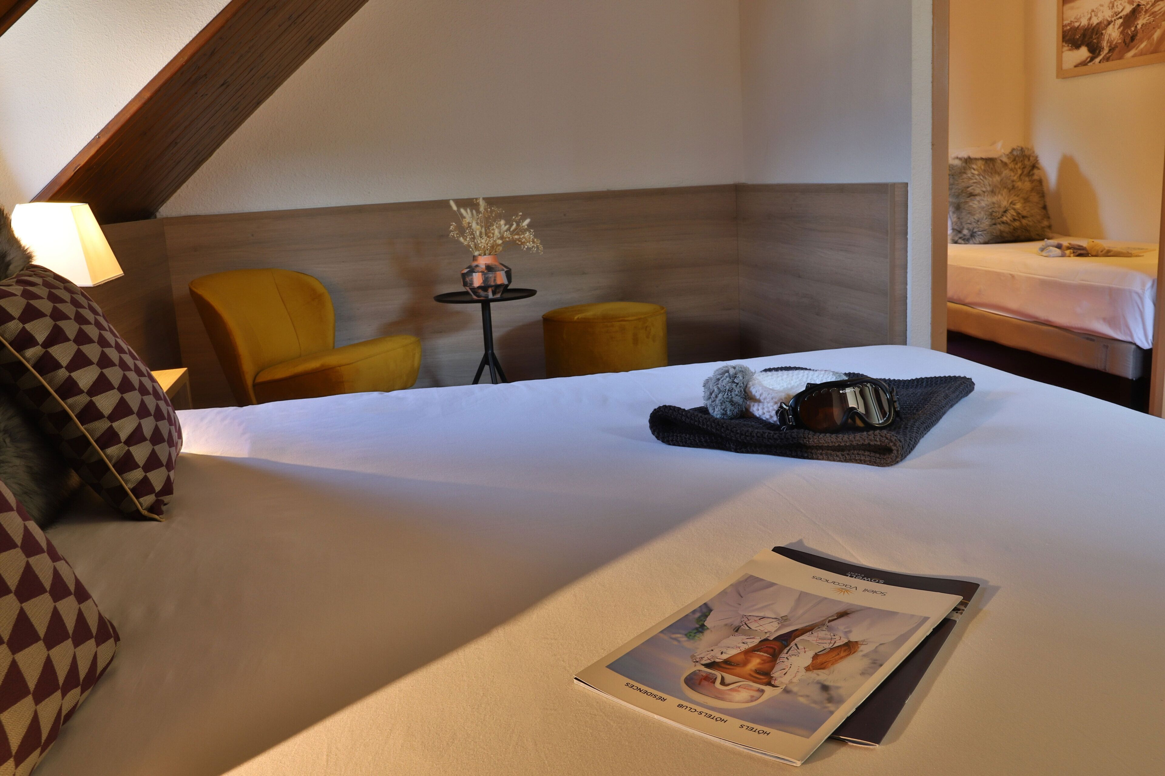 Een hotelkamer met een bed, twee gele fauteuils, een tafeltje met een vaas en een ingelijst foto aan de muur.