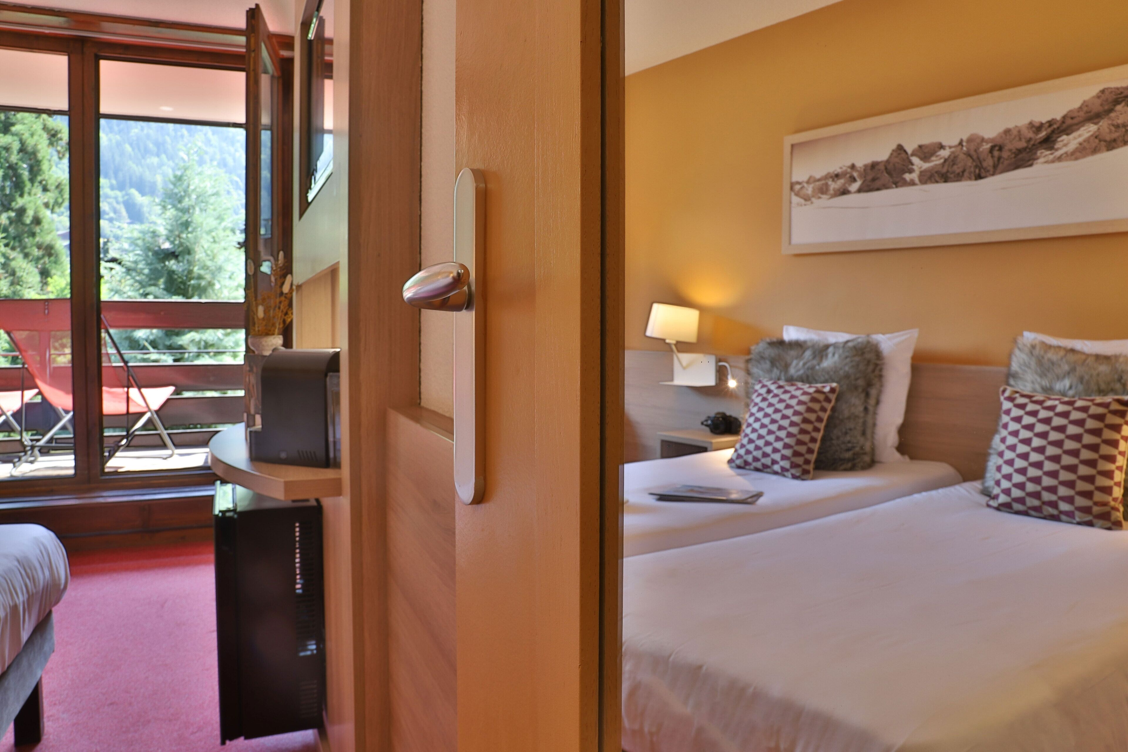 A hotel room with a bed, a desk, a chair, a TV, and a window with a view of trees and mountains.