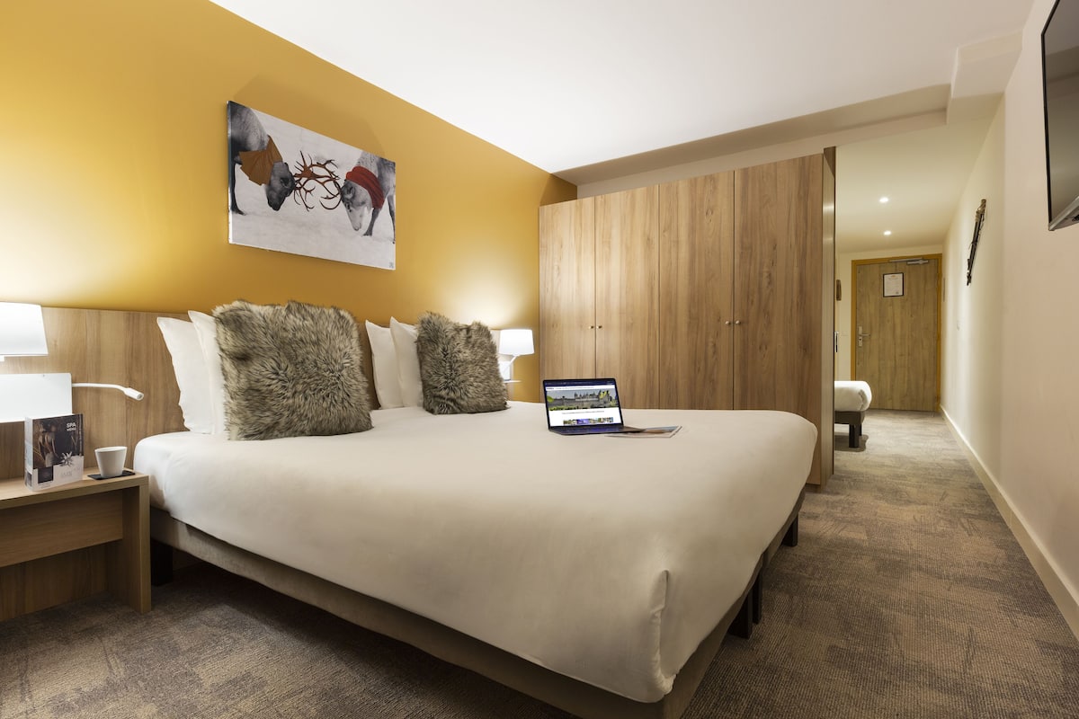 Ein Hotelzimmer mit einem großen Bett, einem Laptop auf dem Bett, einem Nachttisch mit Lampe und einem Gemälde an der Wand.