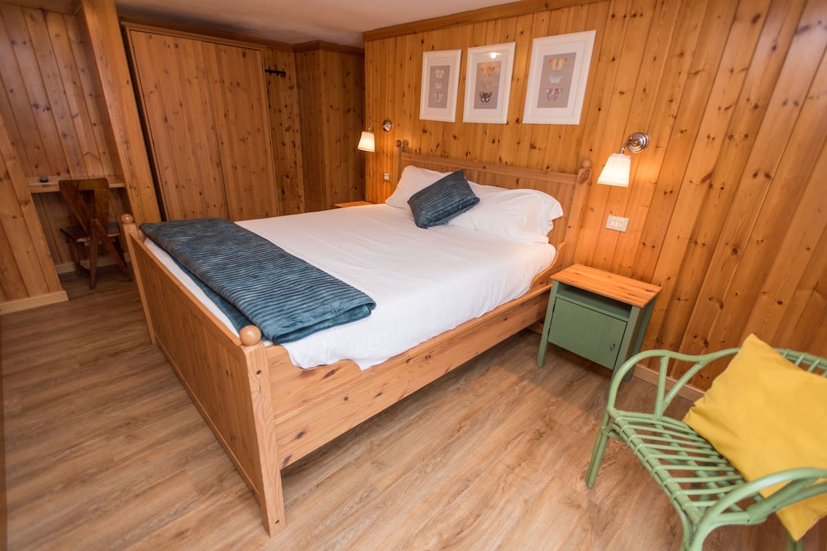 A wooden bed with a white mattress, two pillows, and a blue blanket. There is a green nightstand with a lamp and a chair with a yellow cushion.